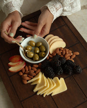 A styled Catholic charcuterie board with sliced cheese, almonds, blackberries, apple slices, and green olives in a small white bowl held by hands wearing a white lace-sleeve top, set on a dark wooden board.