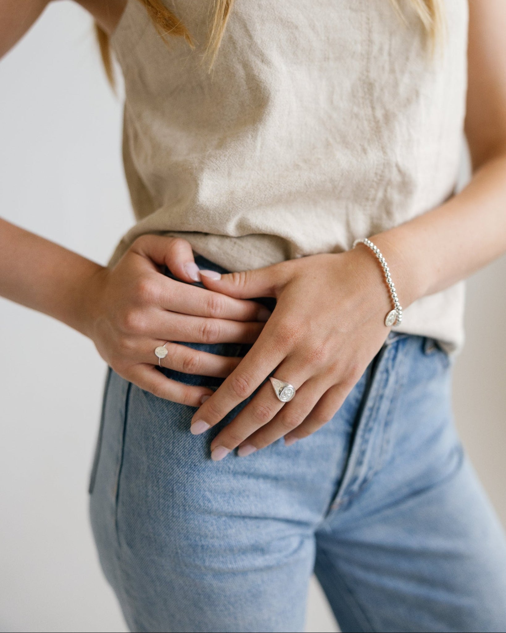 A close-up lifestyle shot of a woman wearing a beige linen tank top and light blue jeans, showcasing a delicate silver chain bracelet and rings on her wrist and fingers.