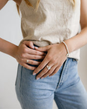 A close-up lifestyle shot of a woman wearing a beige linen tank top and light blue jeans, showcasing a delicate silver chain bracelet and rings on her wrist and fingers.