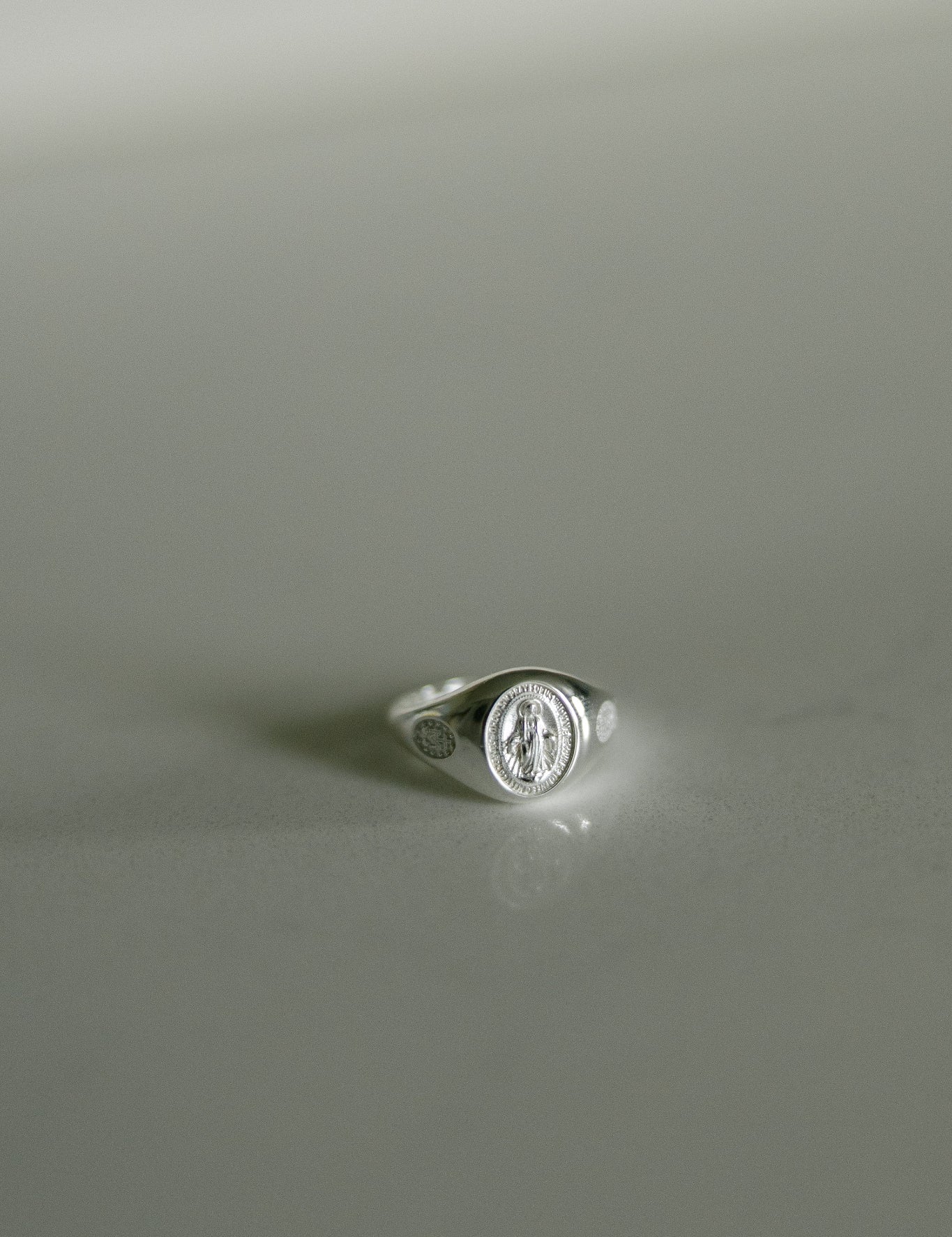 A silver signet ring featuring an oval Miraculous medal on a smooth, light gray reflective surface.