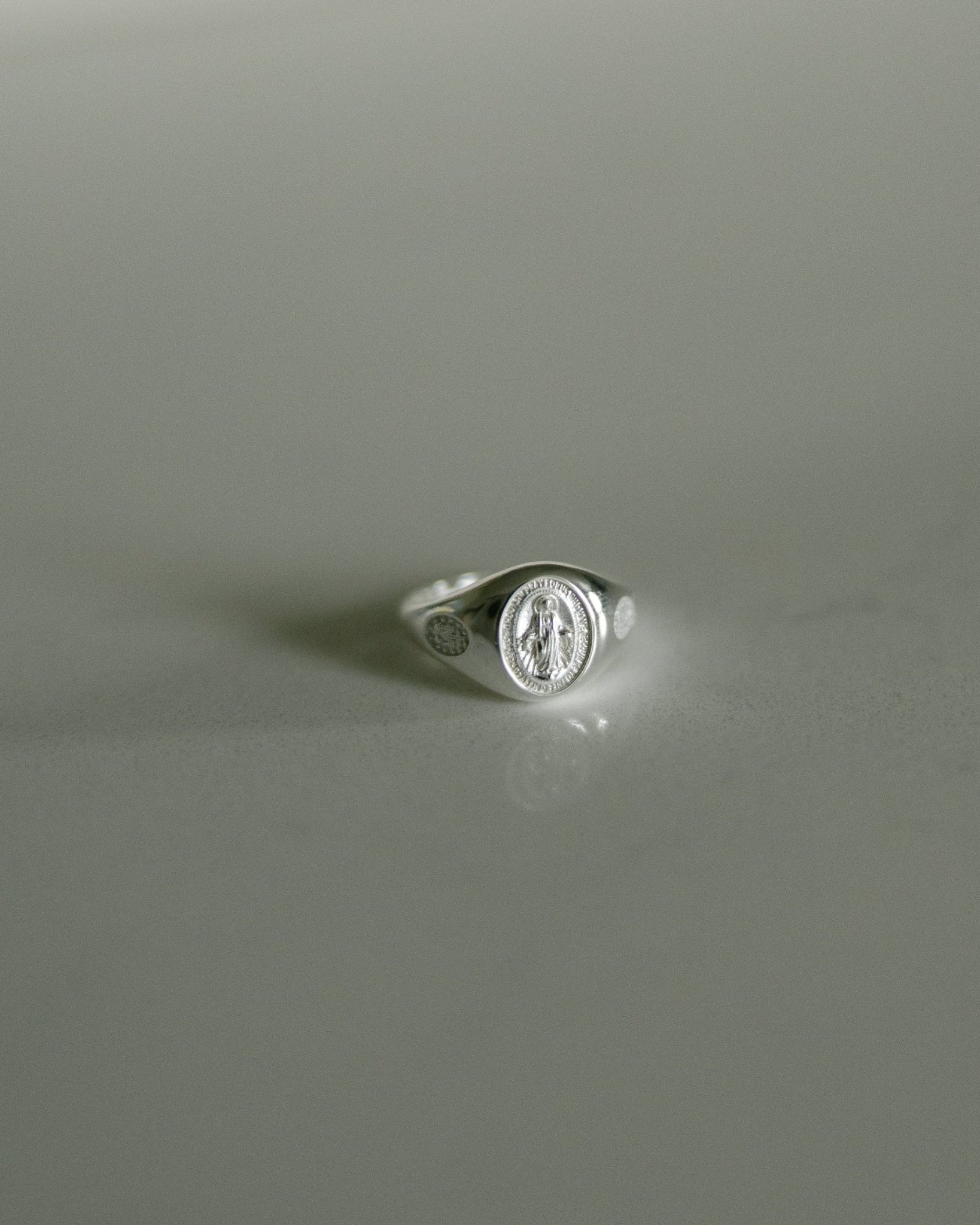 A silver signet ring featuring an oval Miraculous medal on a smooth, light gray reflective surface.