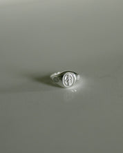 A silver signet ring featuring an oval Miraculous medal on a smooth, light gray reflective surface.