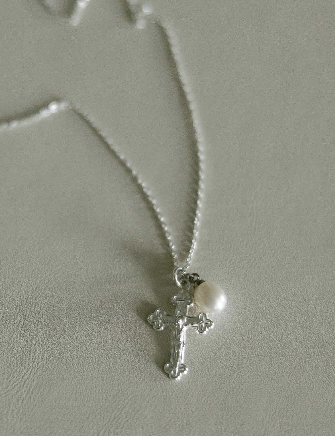 A silver crucifix pendant necklace with a small white pearl, displayed on a textured light gray surface.