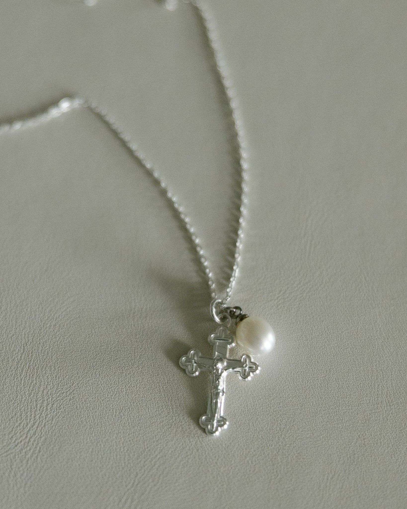 A silver crucifix pendant necklace with a small white pearl, displayed on a textured light gray surface.