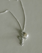 A silver crucifix pendant necklace with a small white pearl, displayed on a textured light gray surface.