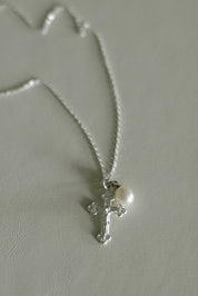 A silver crucifix pendant necklace with a small white pearl, displayed on a textured light gray surface.