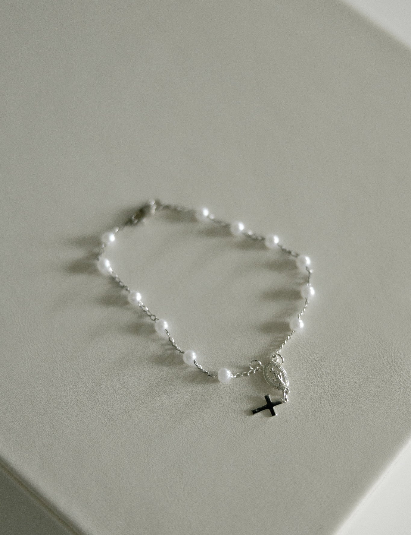 A delicate silver rosary bracelet with small white pearl-like beads and a small black cross pendant, displayed on a light beige textured surface.