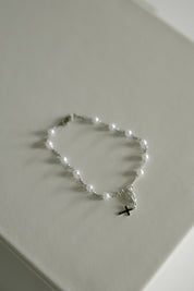 A delicate silver rosary bracelet with small white pearl-like beads and a small black cross pendant, displayed on a light beige textured surface.