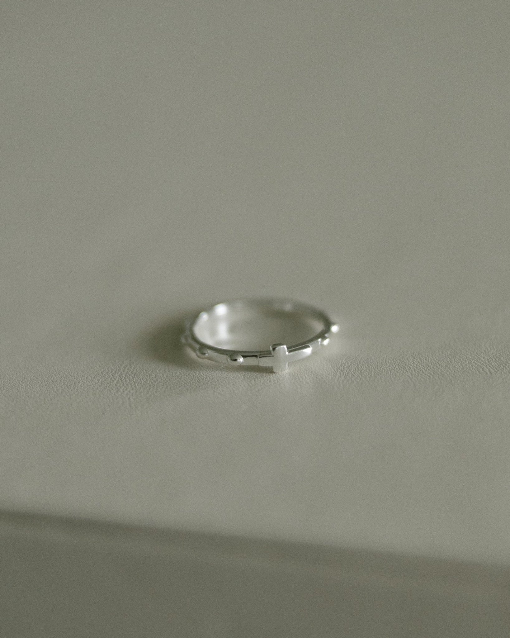 A delicate silver rosary ring resting on a neutral light gray surface.