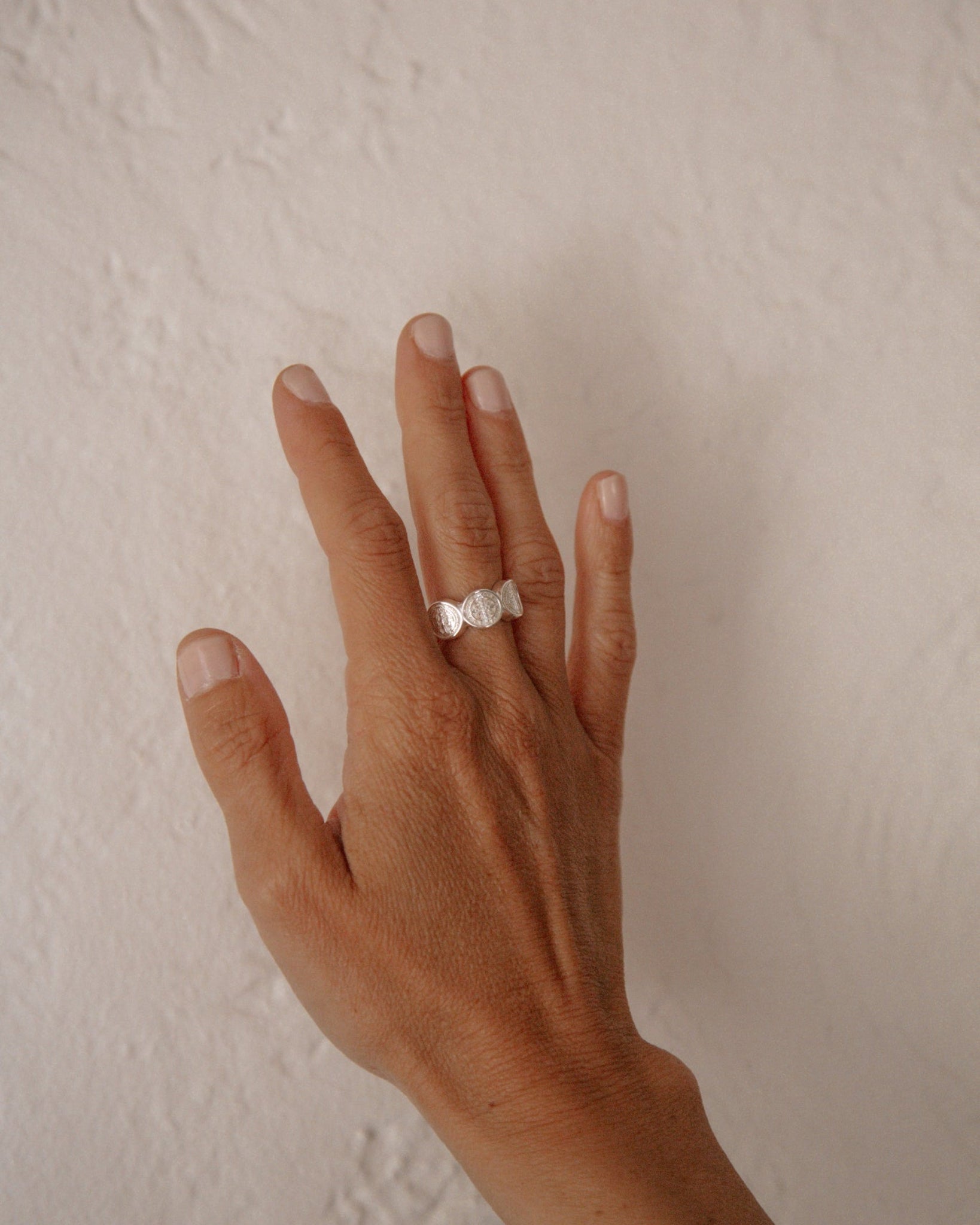 A silver Saint Benedict ring on a person's ring finger, shown against a neutral, textured off-white wall background.
