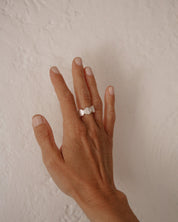 A silver Saint Benedict ring on a person's ring finger, shown against a neutral, textured off-white wall background.