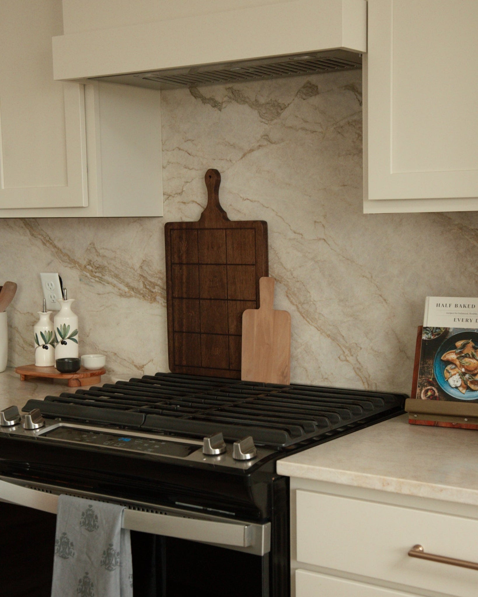 A black gas range stove with a gray patterned towel hanging from the oven handle, set in a kitchen with white cabinets, a beige marble backsplash, West Coast Catholic gospel wooden cutting boards and cookbooks.