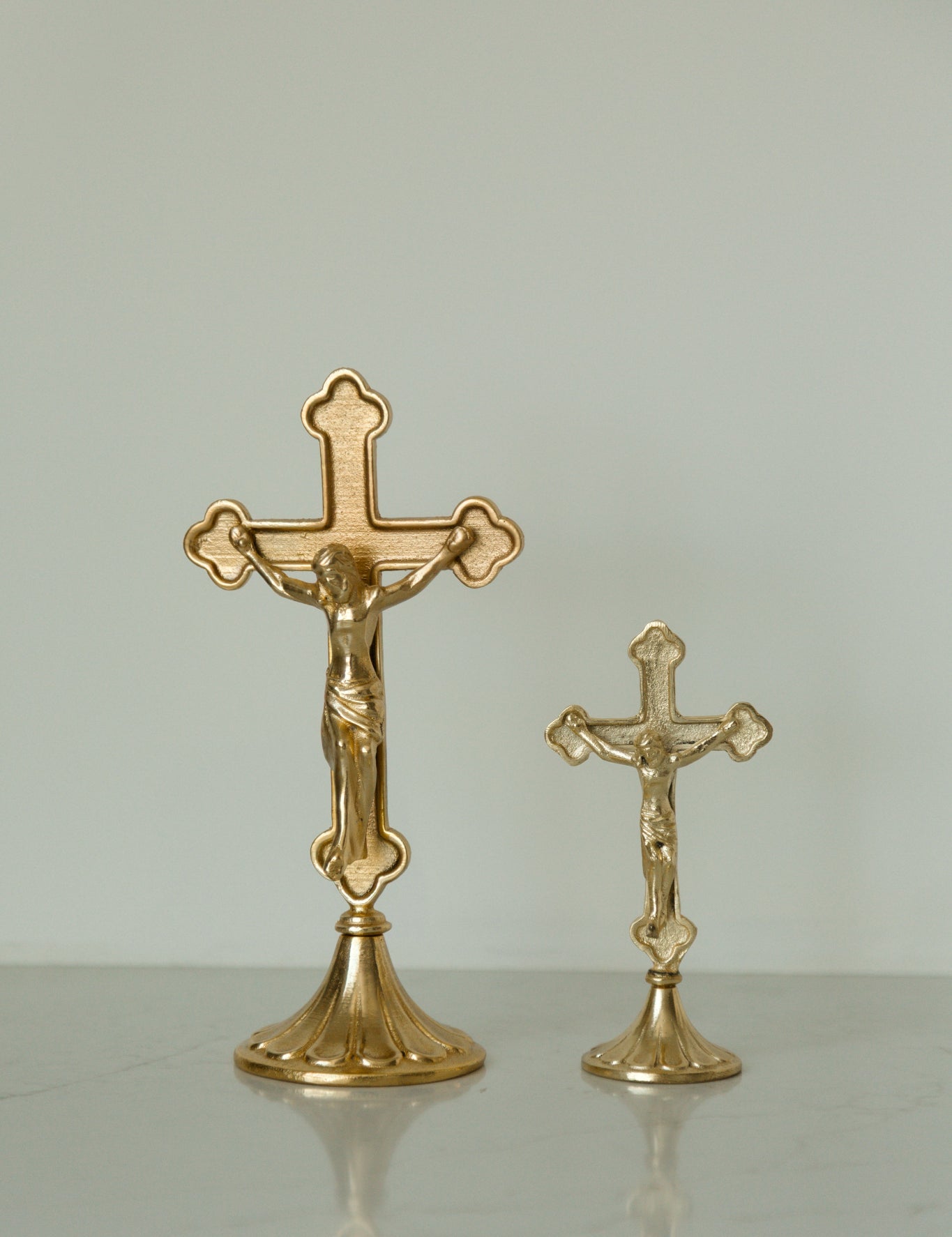 Two gold-toned crucifix home decor on a light gray reflective surface against a neutral light gray background.