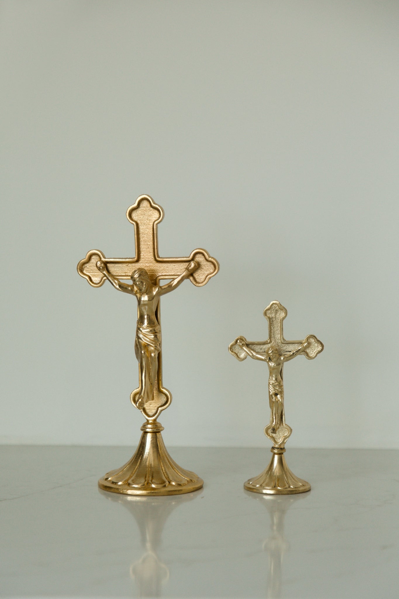 Two gold-toned crucifix home decor on a light gray reflective surface against a neutral light gray background.