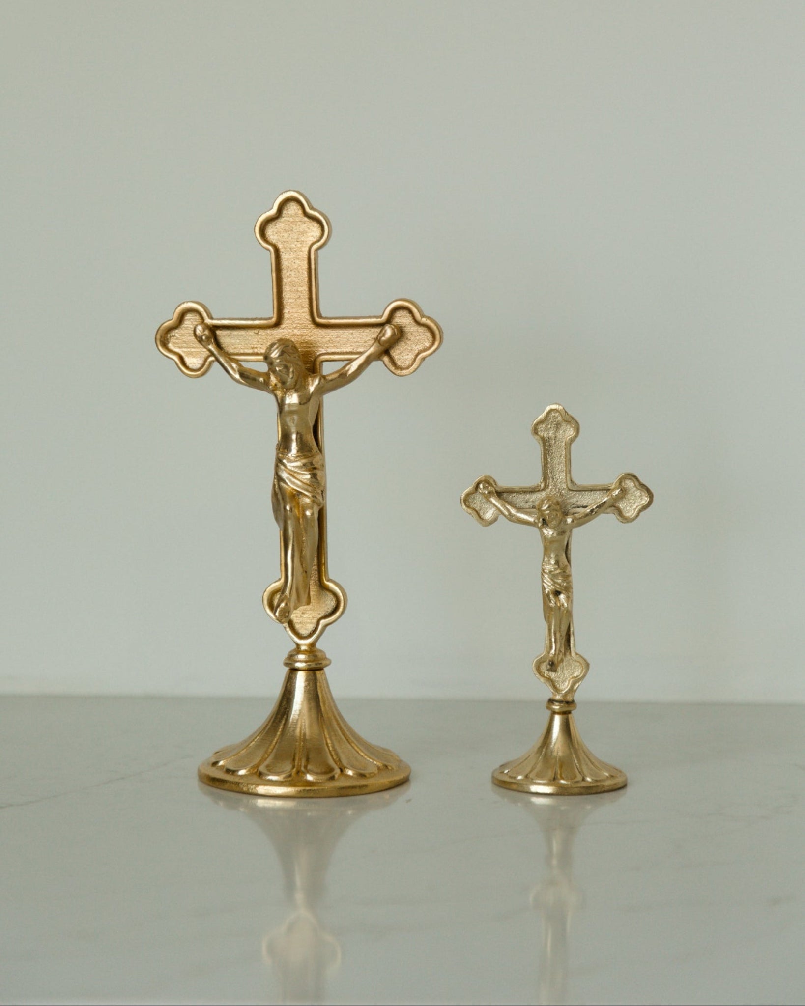 Two gold-toned crucifix home decor on a light gray reflective surface against a neutral light gray background.