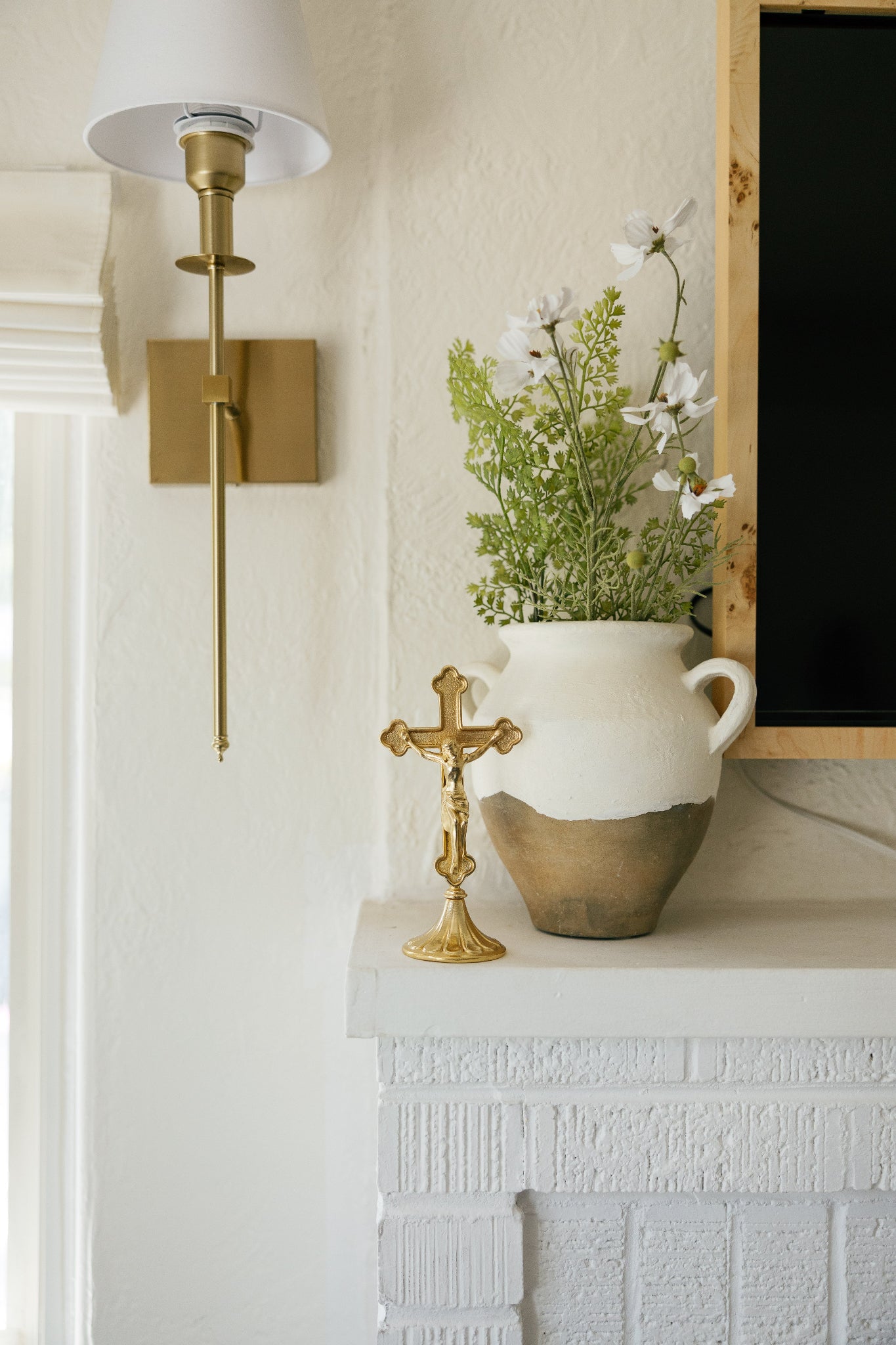 Gold standing crucifix on a white brick fireplace beside a rustic vase of white flowers, modern Catholic home decor.