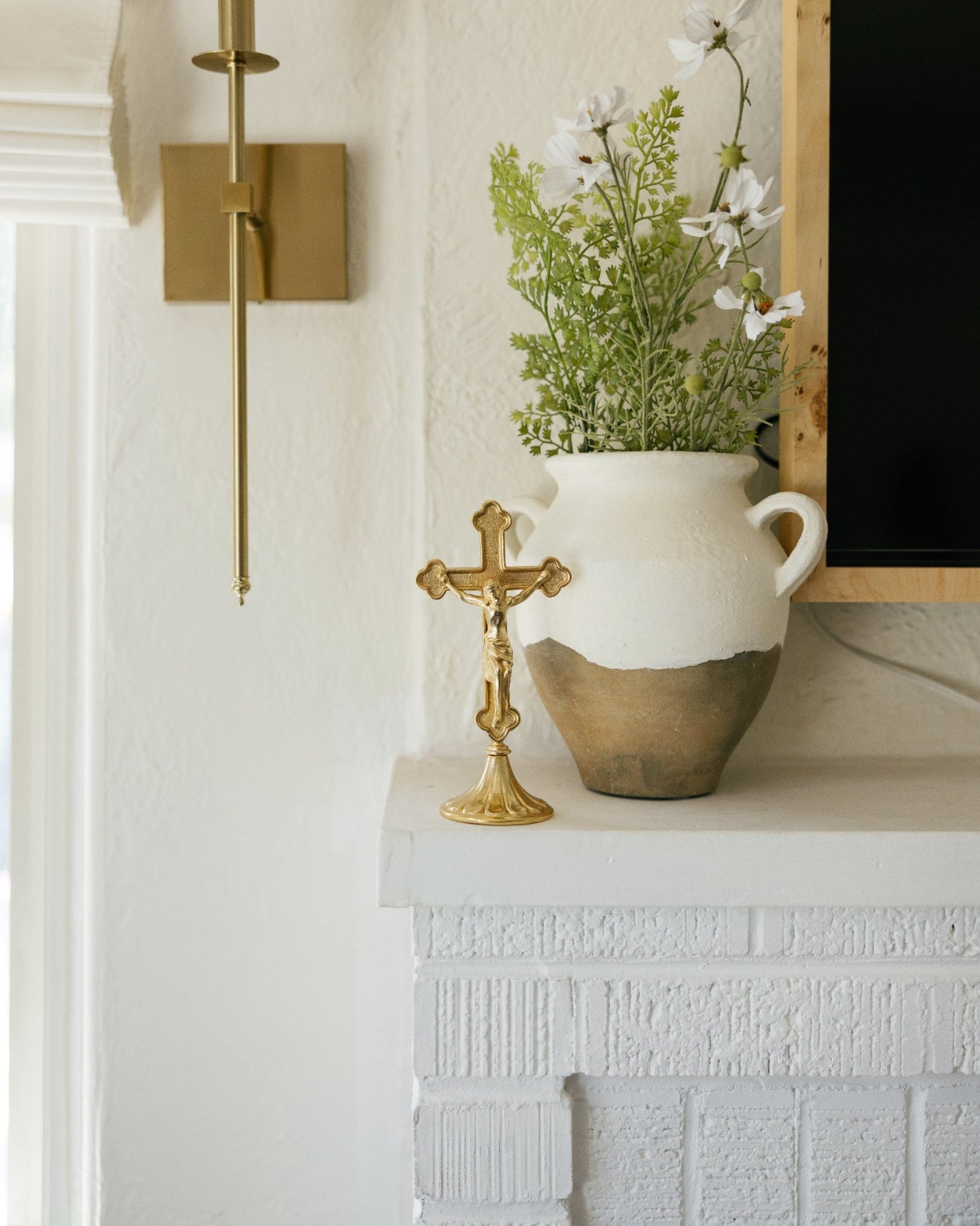 Gold standing crucifix on a white brick fireplace beside a rustic vase of white flowers, modern Catholic home decor.