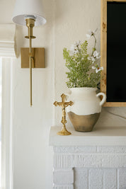 Gold standing crucifix on a white brick fireplace beside a rustic vase of white flowers, modern Catholic home decor.