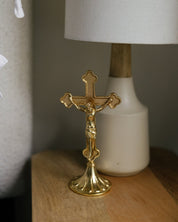 A small gold standing crucifix on a wooden nightstand beside a linen lamp, perfect for modern Catholic home decor for a prayer corner