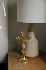 A small gold standing crucifix on a wooden nightstand beside a linen lamp, perfect for modern Catholic home decor for a prayer corner