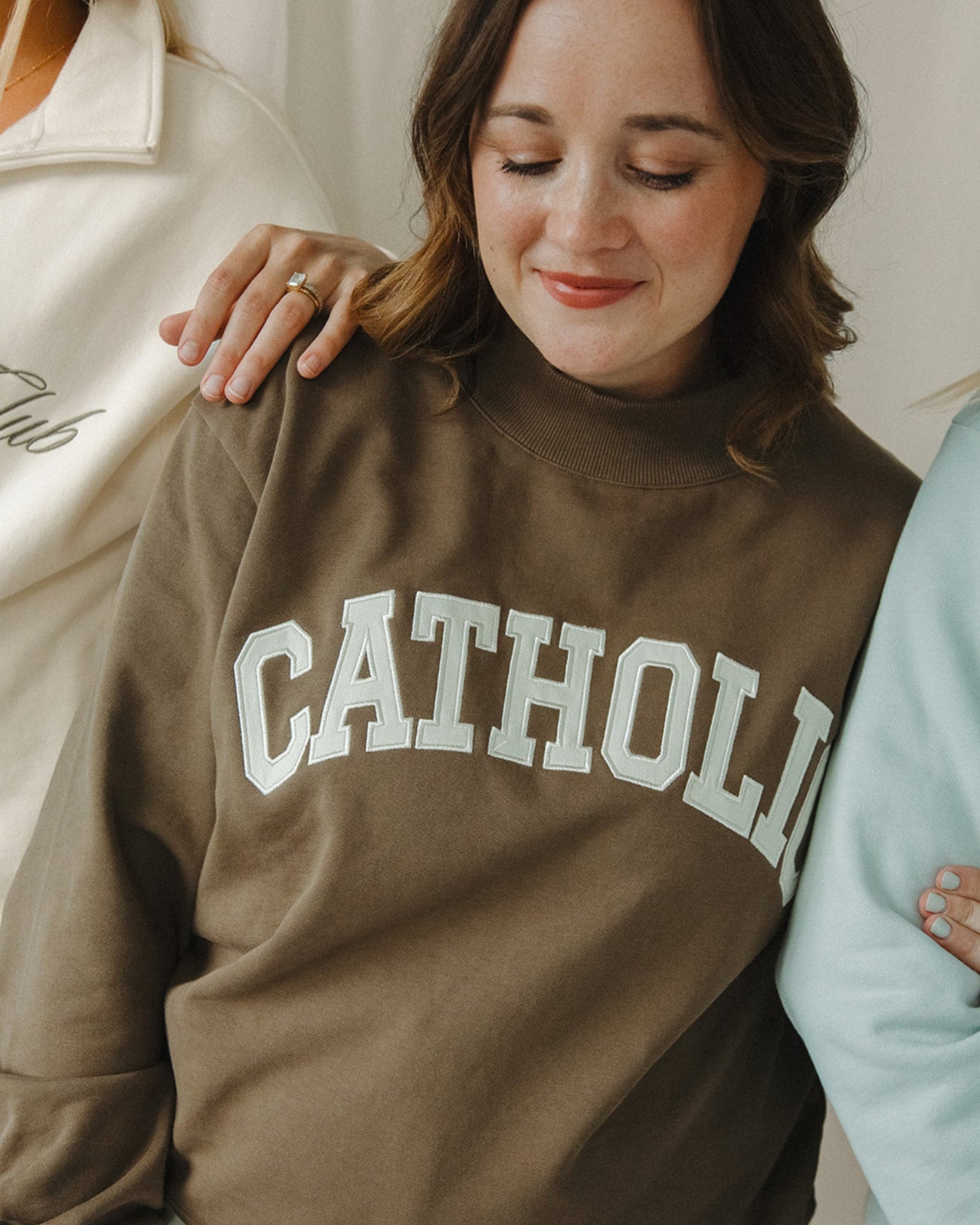 Woman wearing a brown sweatshirt with 'CATHOLIC' printed on it, standing between two other people.