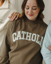 Woman wearing a brown sweatshirt with 'CATHOLIC' printed on it, standing between two other people.