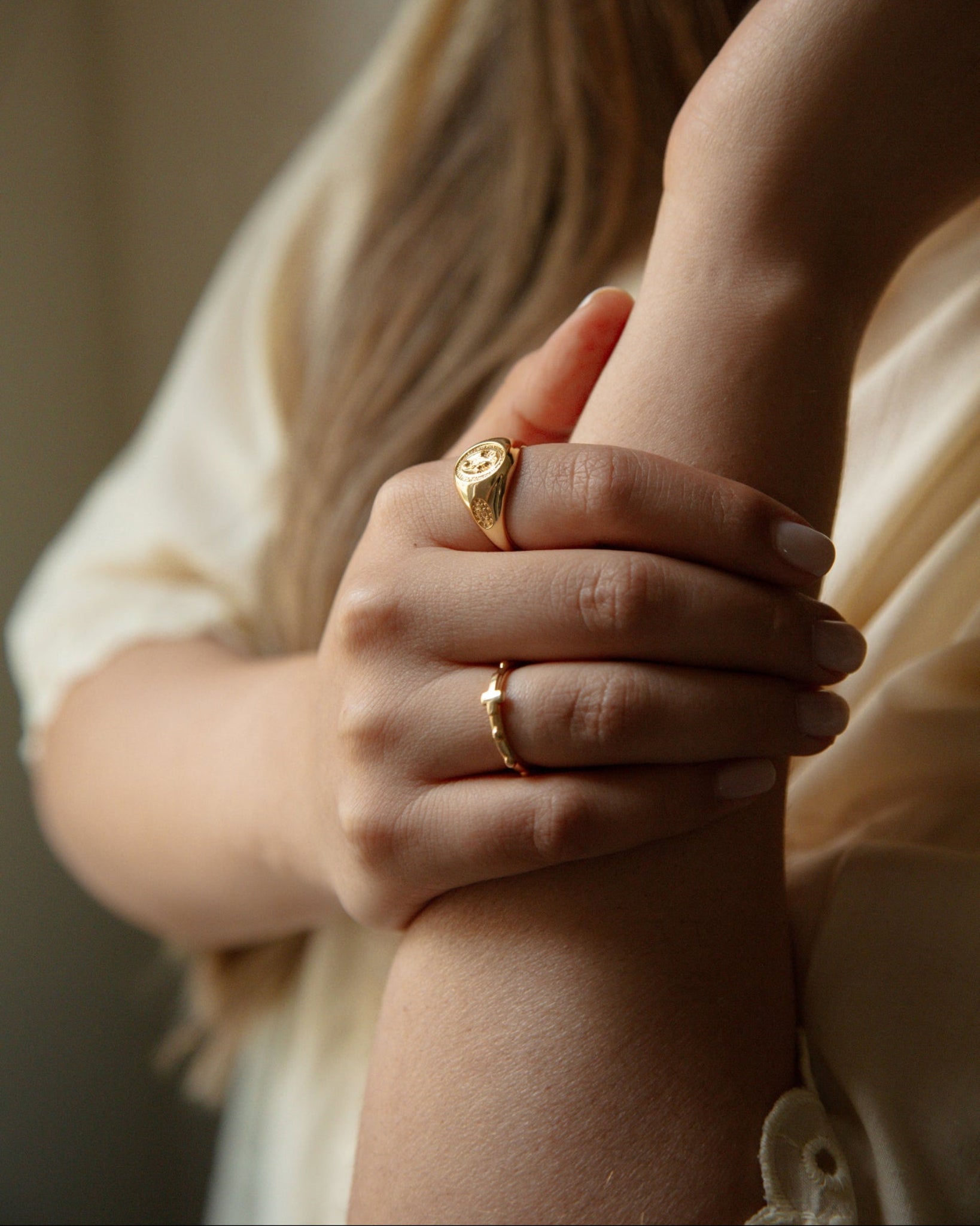 A close-up lifestyle shot of a woman wearing two catholic gold rings on her fingers, with her hand resting on her arm. She is wearing a cream-colored blouse, and the background is a soft, warm-toned neutral indoor setting.