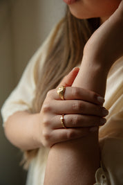 A close-up lifestyle shot of a woman wearing two catholic gold rings on her fingers, with her hand resting on her arm. She is wearing a cream-colored blouse, and the background is a soft, warm-toned neutral indoor setting.