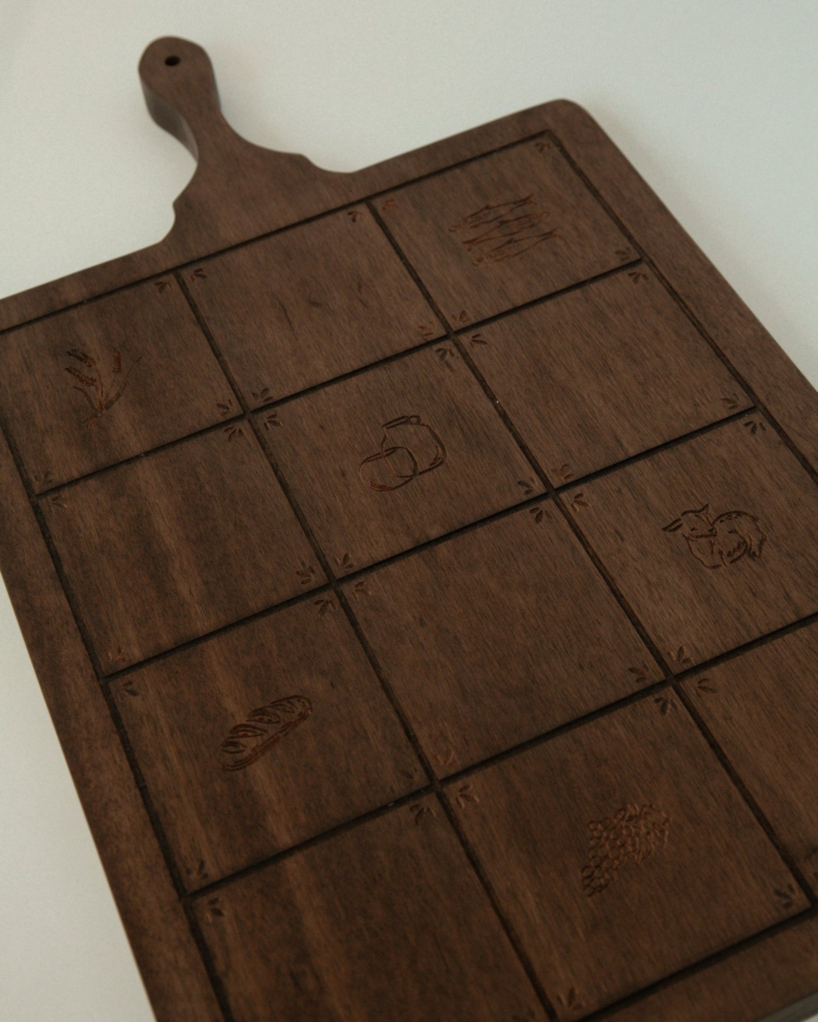 A dark brown wooden Catholic serving board with a handle, featuring a 3x3 grid of square compartments with engraved food icons, photographed on a light off-white background.