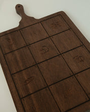 A dark brown wooden Catholic serving board with a handle, featuring a 3x3 grid of square compartments with engraved food icons, photographed on a light off-white background.