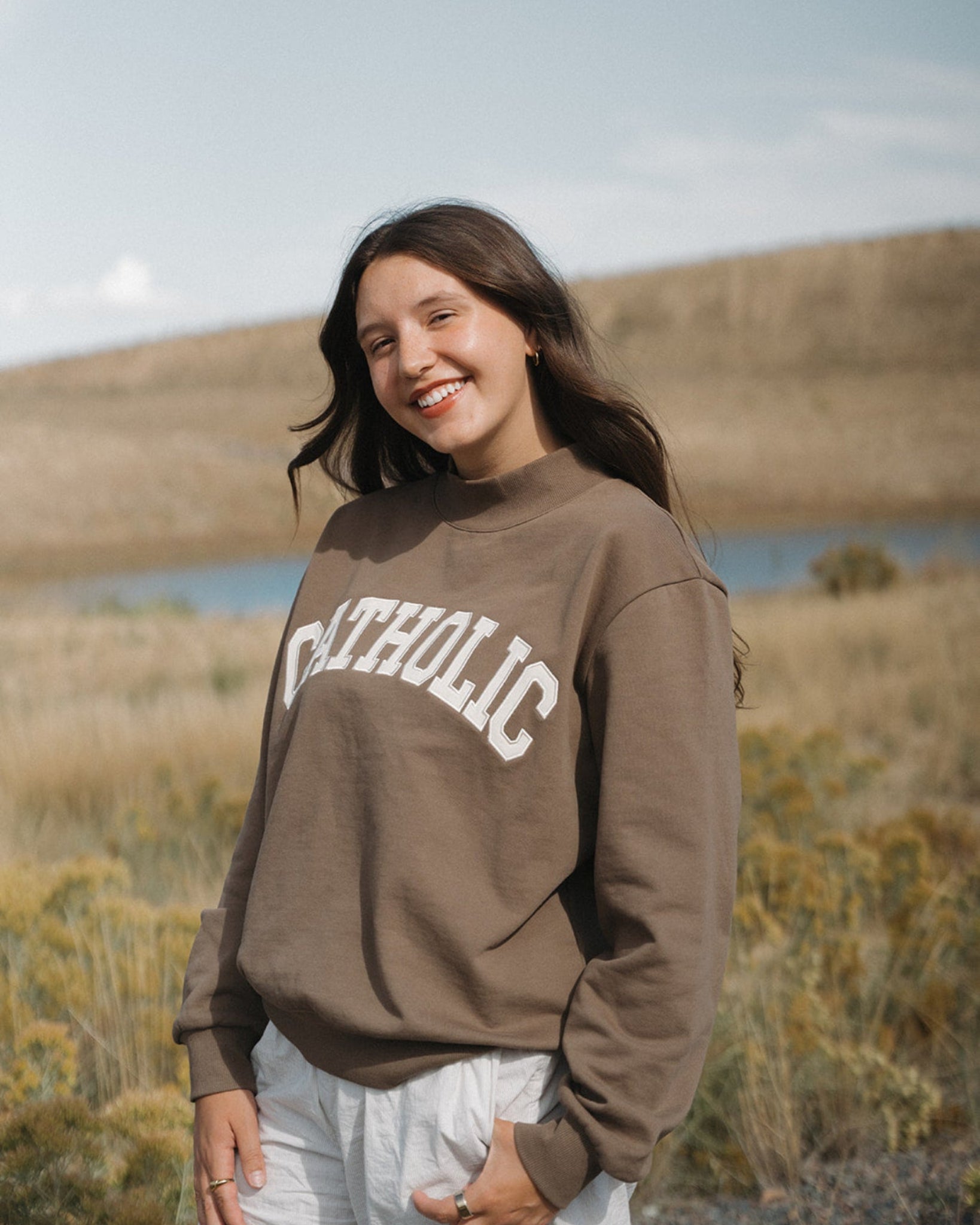 Woman wearing a brown sweatshirt with 'Catholic' text, standing in a scenic outdoor setting.