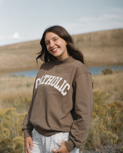 Woman wearing a brown sweatshirt with 'Catholic' text, standing in a scenic outdoor setting.