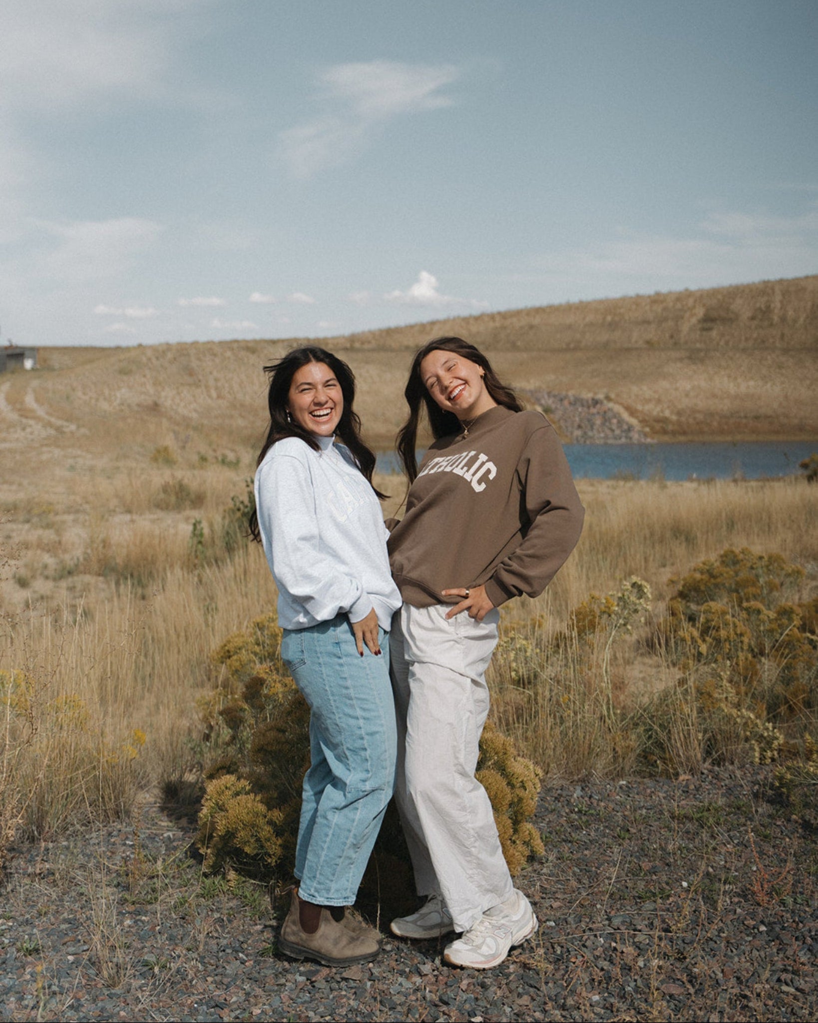 Two women standing outdoors in a grassy field near a lake under a blue sky, wearing casual sweatshirts and jeans. One sweatshirt has a visible 'SUNRISE' graphic.