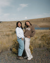 Two women standing outdoors in a grassy field near a lake under a blue sky, wearing casual sweatshirts and jeans. One sweatshirt has a visible 'SUNRISE' graphic.