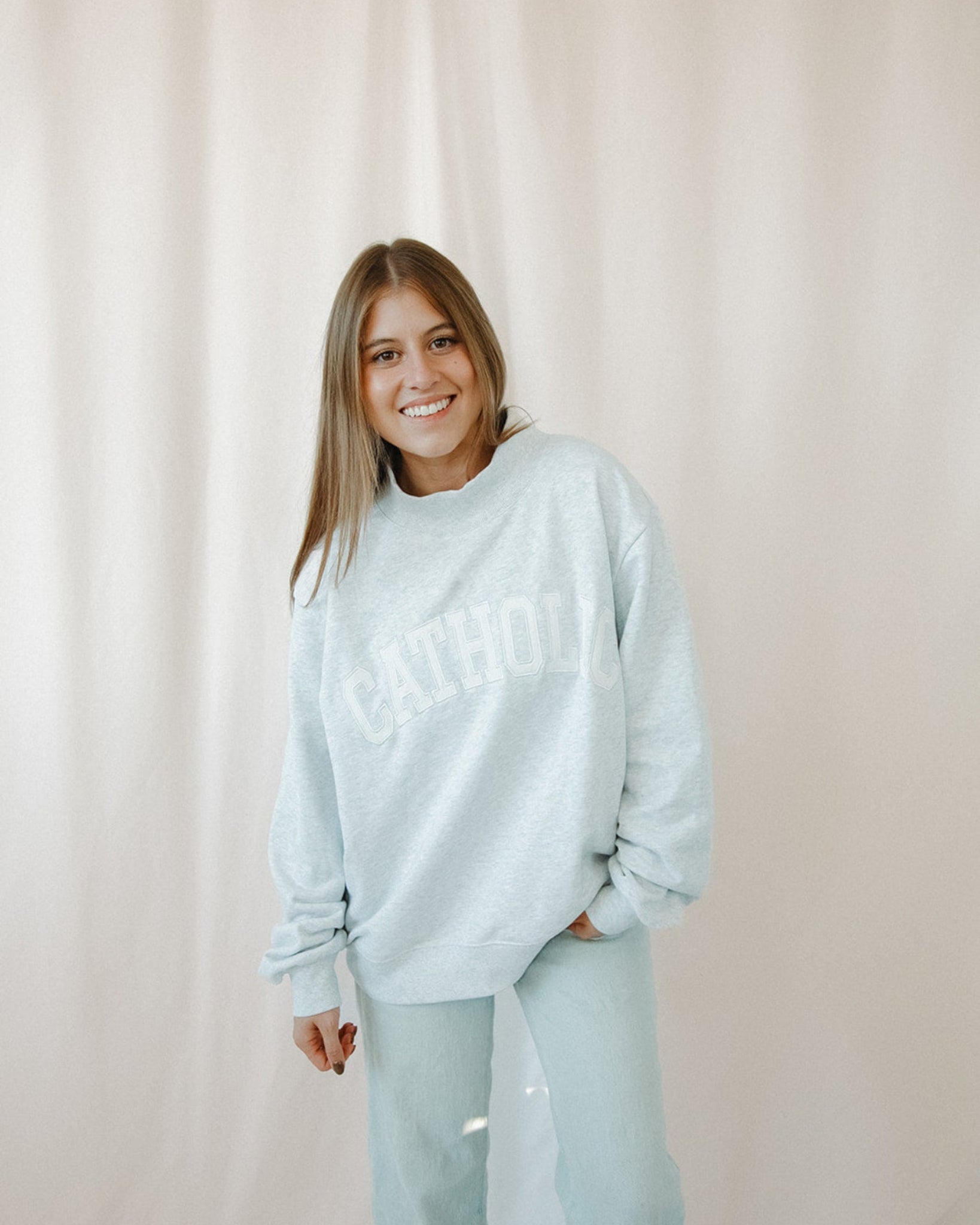 Woman wearing a light grey catholic mockneck sweatshirt and pants against a beige curtain background
