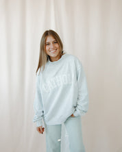 Woman wearing a light grey catholic mockneck sweatshirt and pants against a beige curtain background