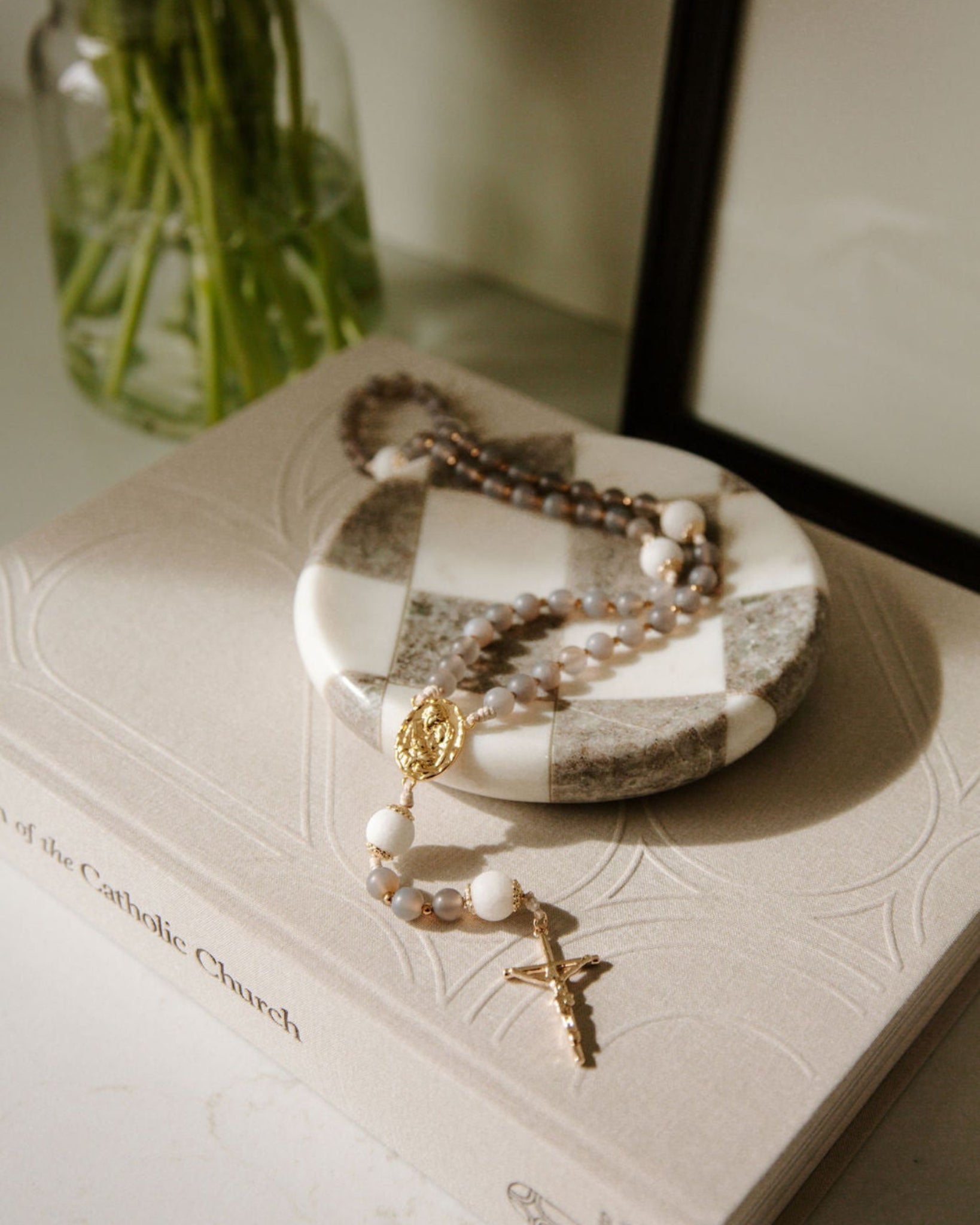 West Coast Catholic, Holy Family rosary on a checkered dish with a coffee table book underneath, next to a vase with greenery.