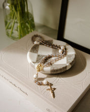 West Coast Catholic, Holy Family rosary on a checkered dish with a coffee table book underneath, next to a vase with greenery.