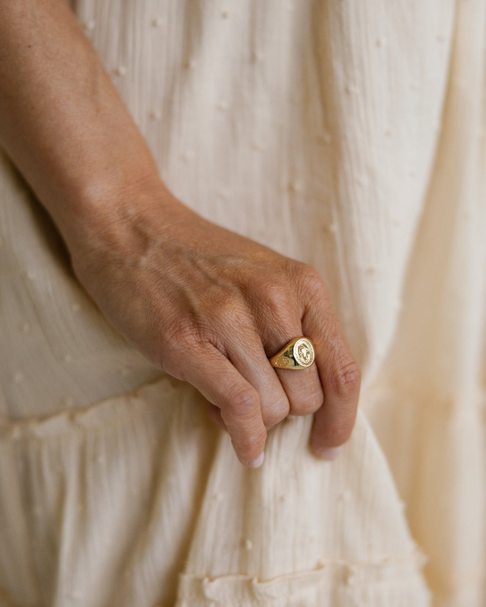 A close-up of a person's hand wearing a gold Miraculous medal signet ring set against a cream-colored textured dress.