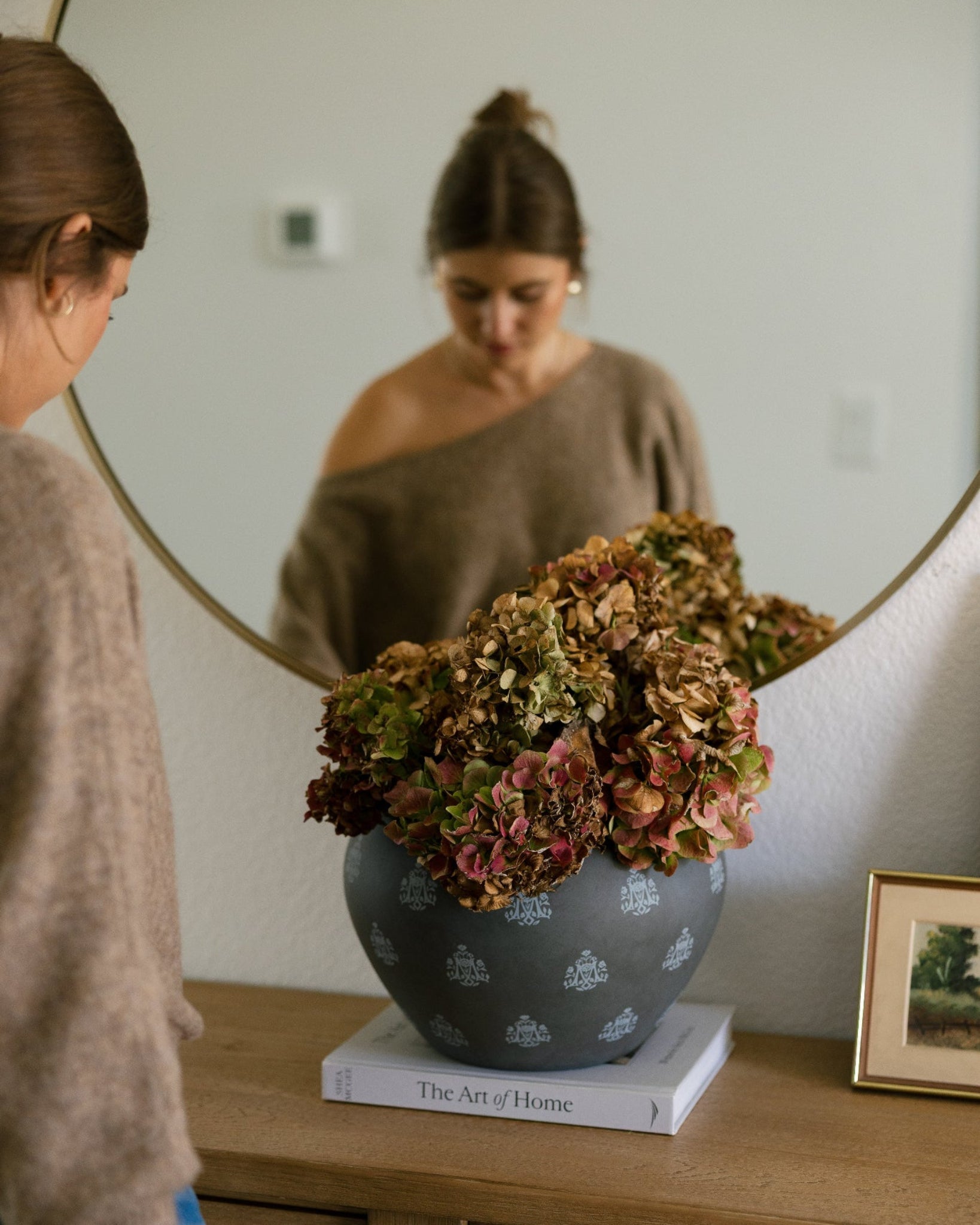 A woman in a beige sweater stands beside a wooden dresser, looking at her reflection in a large oval mirror. The reflection shows her wearing a taupe one-shoulder top.
