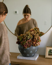 A woman in a beige sweater stands beside a wooden dresser, looking at her reflection in a large oval mirror. The reflection shows her wearing a taupe one-shoulder top.