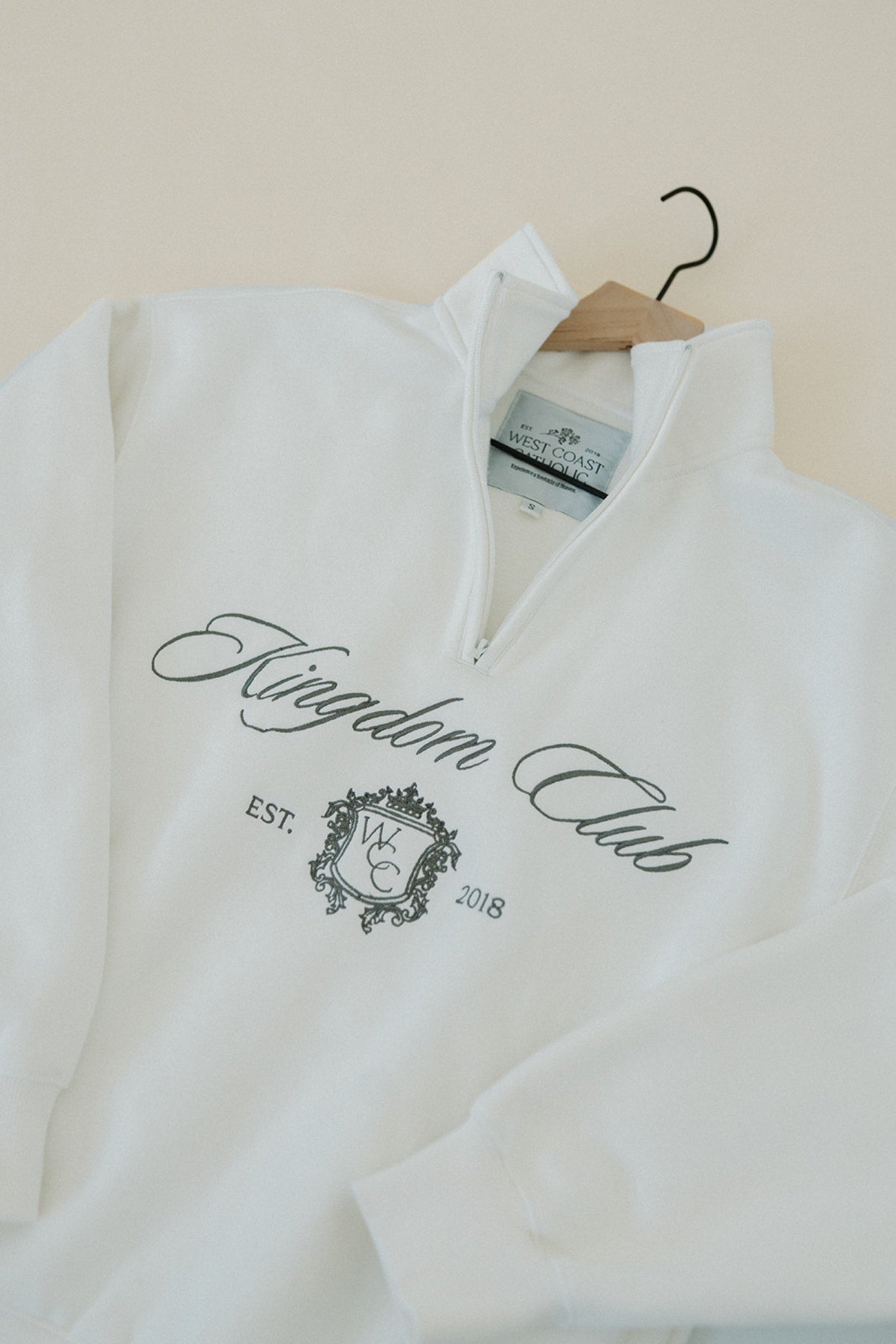 White sweatshirt with 'Kingdom Club' text and logo on a hanger against a beige wall.