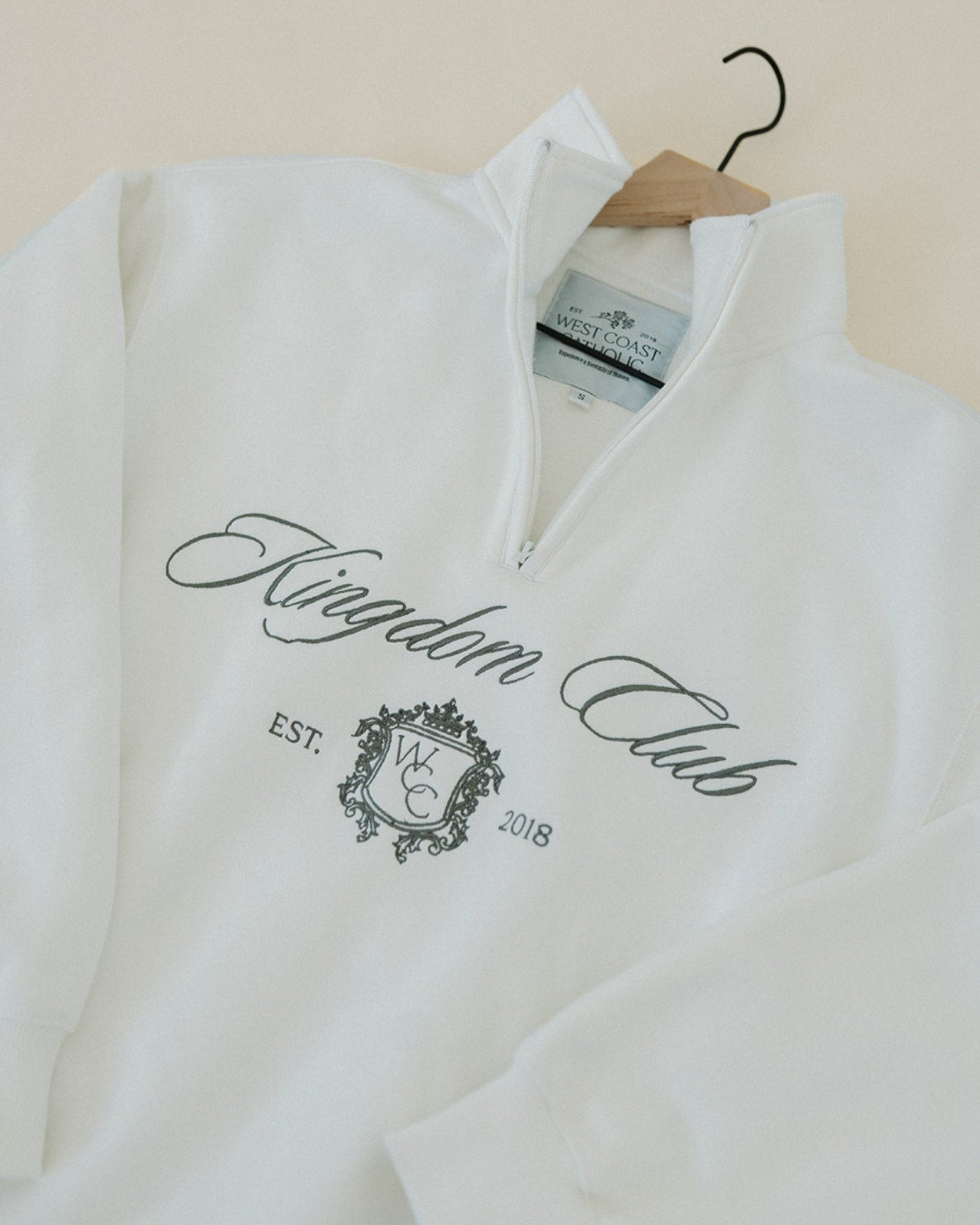 White sweatshirt with 'Kingdom Club' text and logo on a hanger against a beige wall.