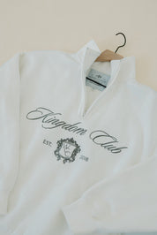 White sweatshirt with 'Kingdom Club' text and logo on a hanger against a beige wall.
