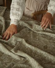 A close-up of a person's hands and torso smoothing out a patterned olive green Catholic tablecloth while wearing a white lace long-sleeve top and a beige apron.
