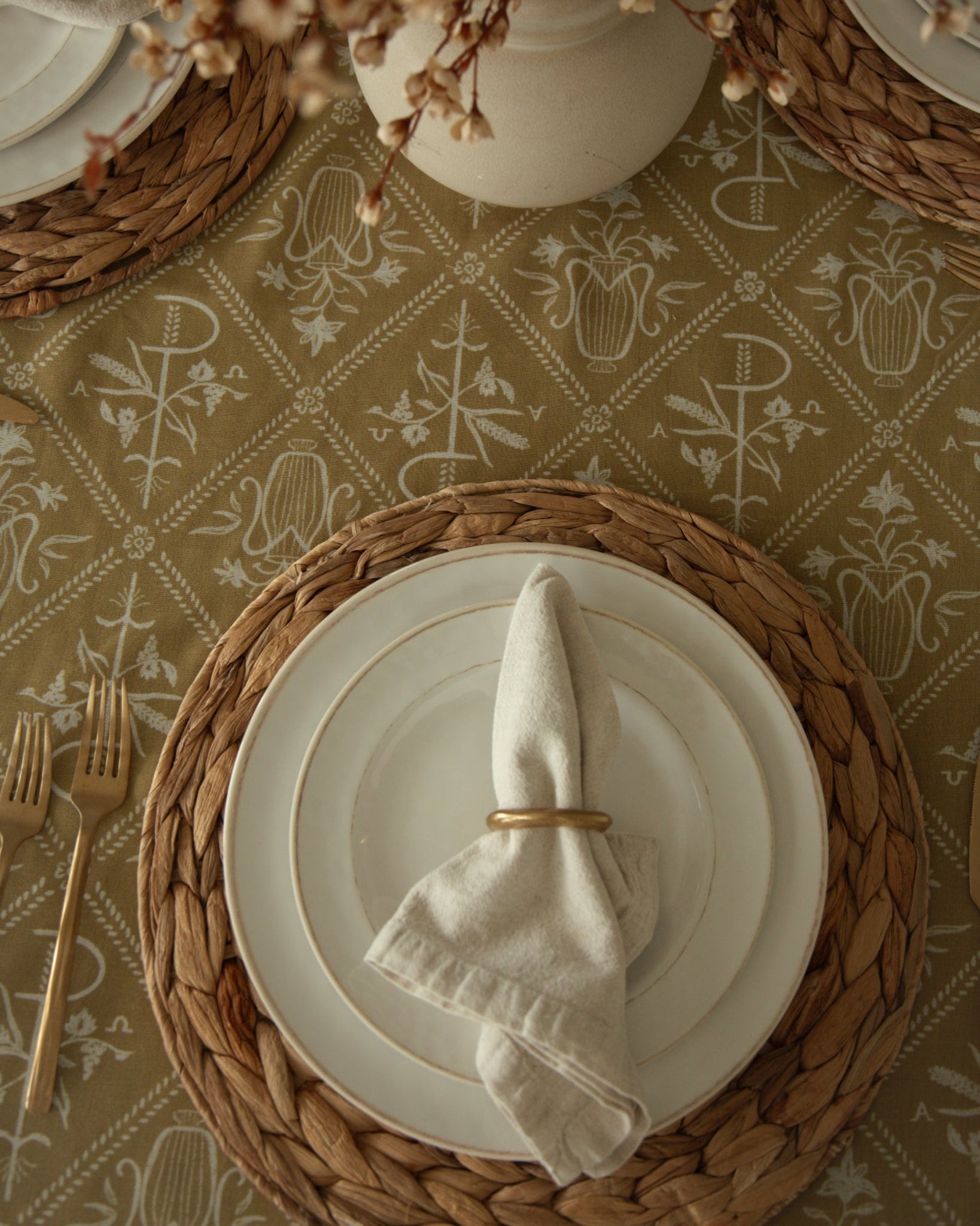A styled tabletop setting with stacked white plates on woven round placemats, a folded cream napkin with a gold ring, a Catholic inspired mustard yellow tablecloth, and a white vase of dried flowers.