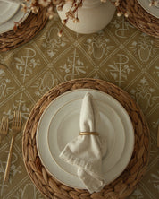 A styled tabletop setting with stacked white plates on woven round placemats, a folded cream napkin with a gold ring, a Catholic inspired mustard yellow tablecloth, and a white vase of dried flowers.