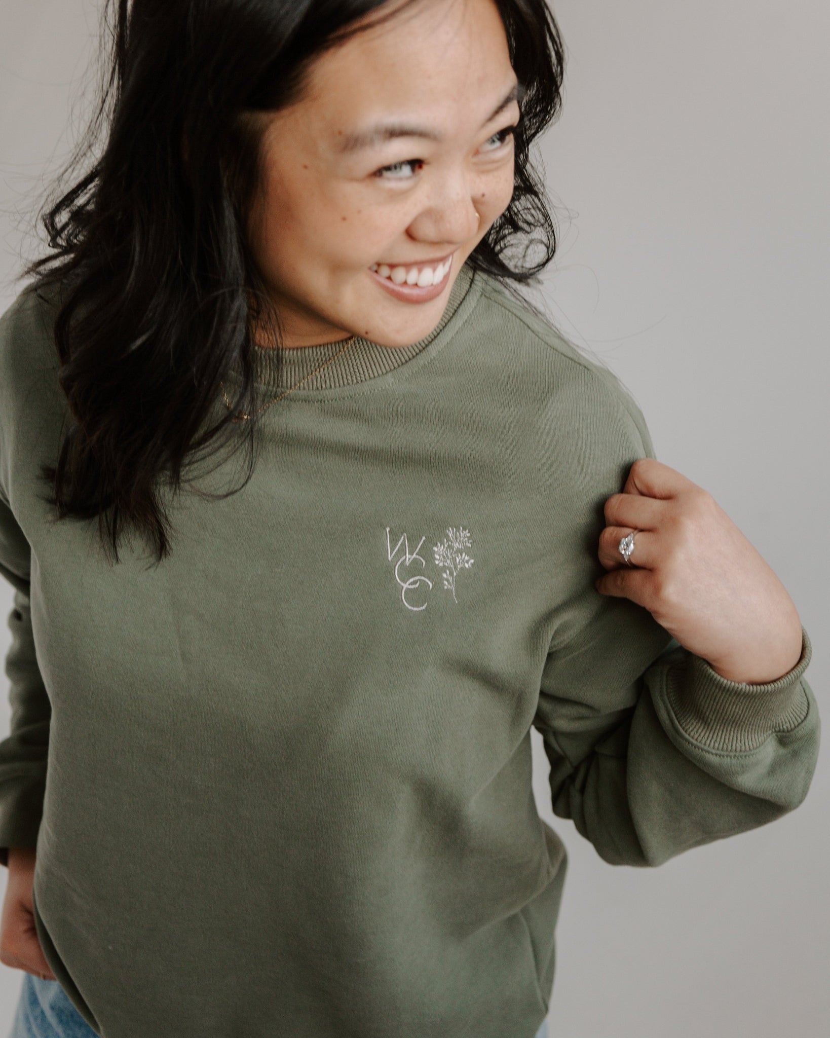 A woman smiling while wearing an olive green crewneck sweatshirt with a logo on the left chest area.