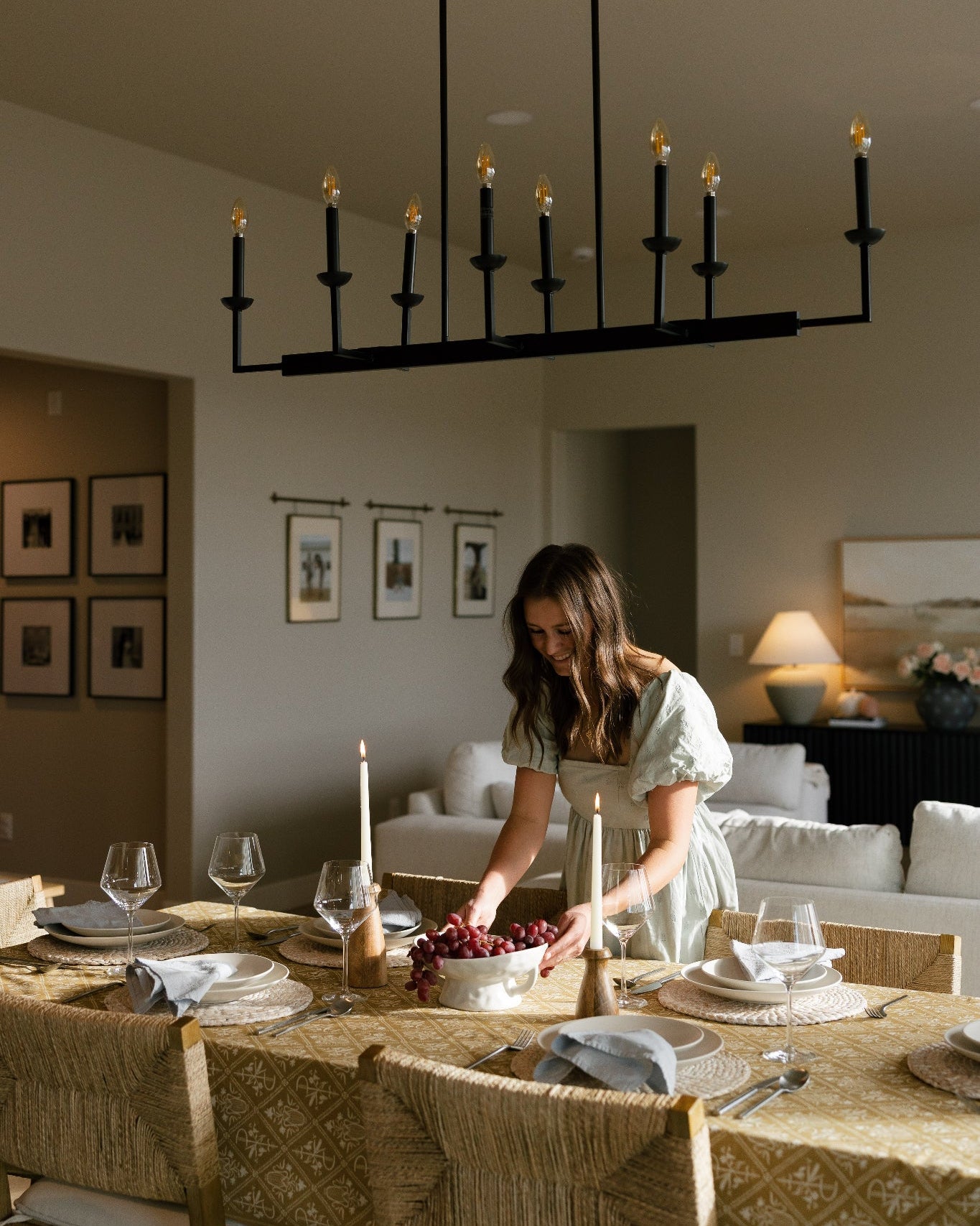 A woman in a light green dress stands at a dining table set for a dinner party, with a large black multi-arm chandelier hanging above. The room has warm neutral walls, framed art, and a cozy living area in the background.