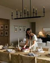 A woman in a light green dress stands at a dining table set for a dinner party, with a large black multi-arm chandelier hanging above. The room has warm neutral walls, framed art, and a cozy living area in the background.